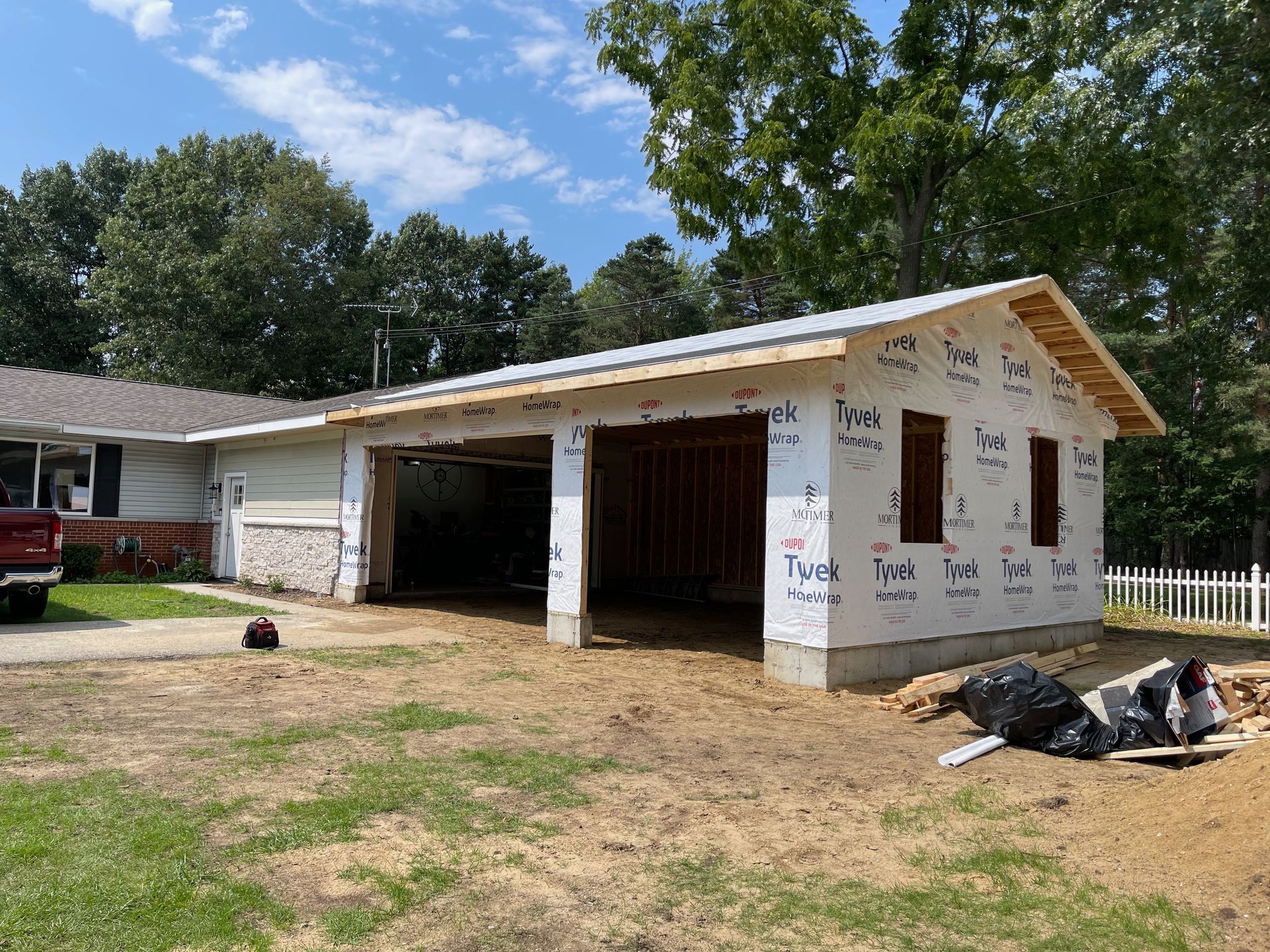 Garage construction next to a house; unfinished exterior, Tyvek wrap, blue sky, and green trees.
