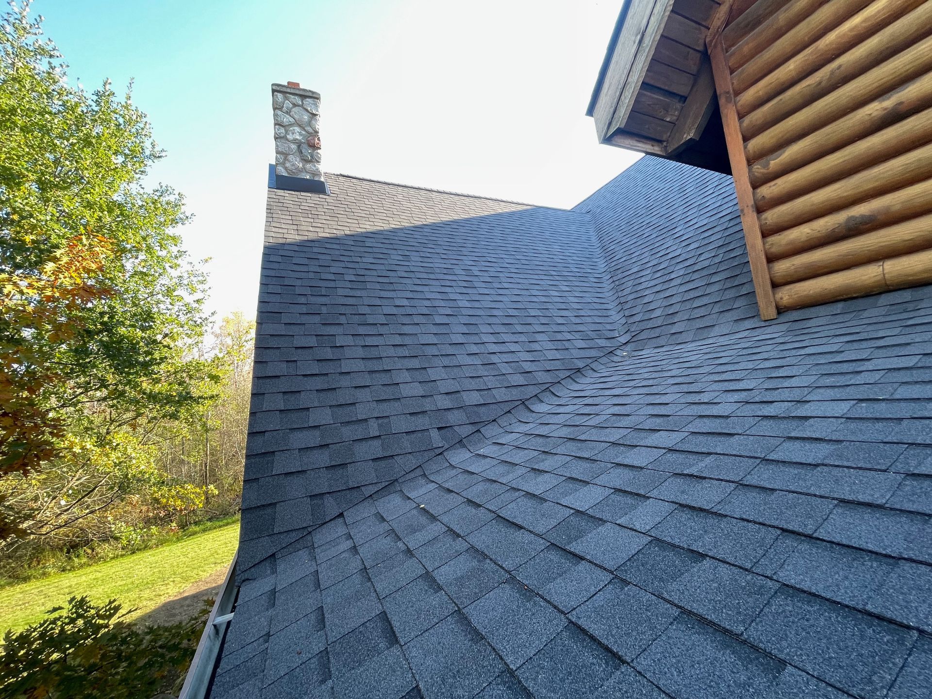 Black shingle roof with a stone chimney, a log cabin wall, and green foliage under a blue sky.