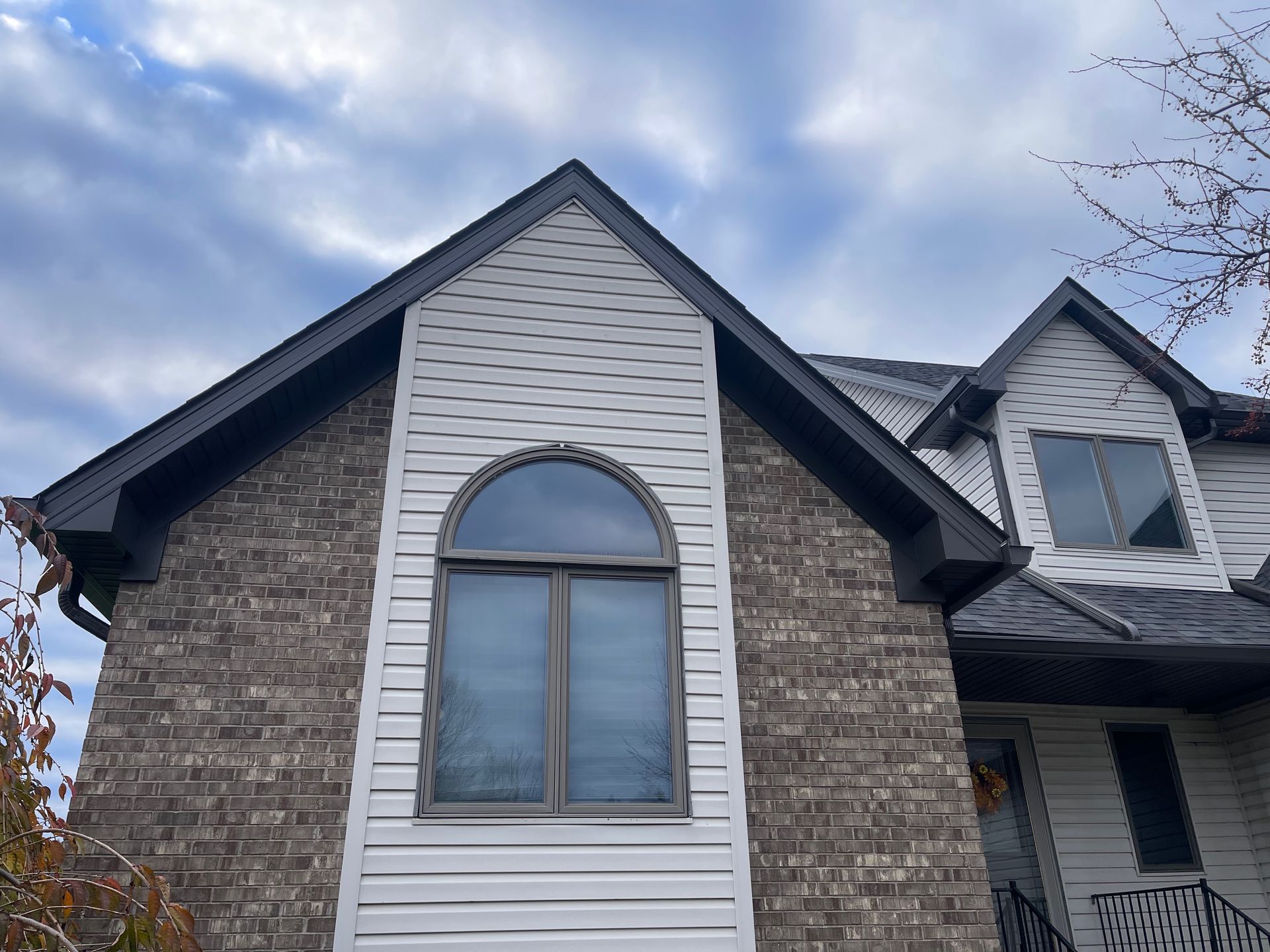 Two-story house with brick and white siding, dark roof, arched window, and overcast sky.