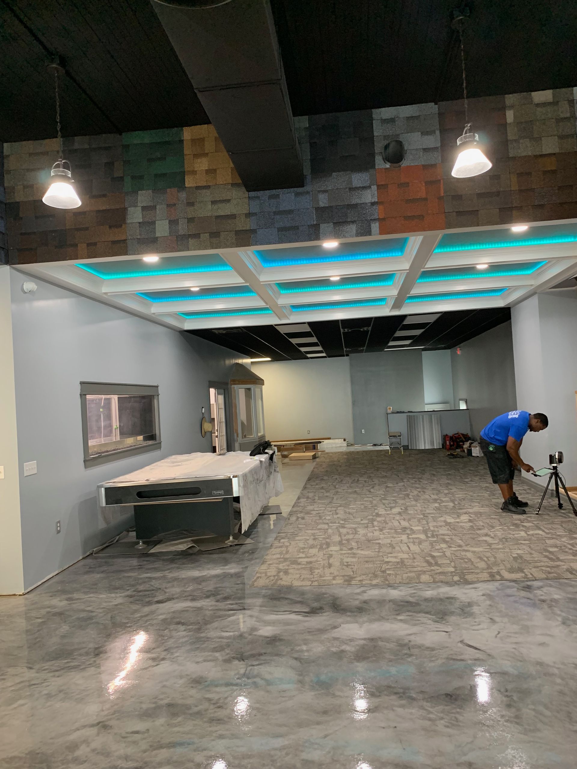 Construction in a commercial space, with exposed ceiling tiles, stone accent wall, gray concrete floor. A worker works on the wall.