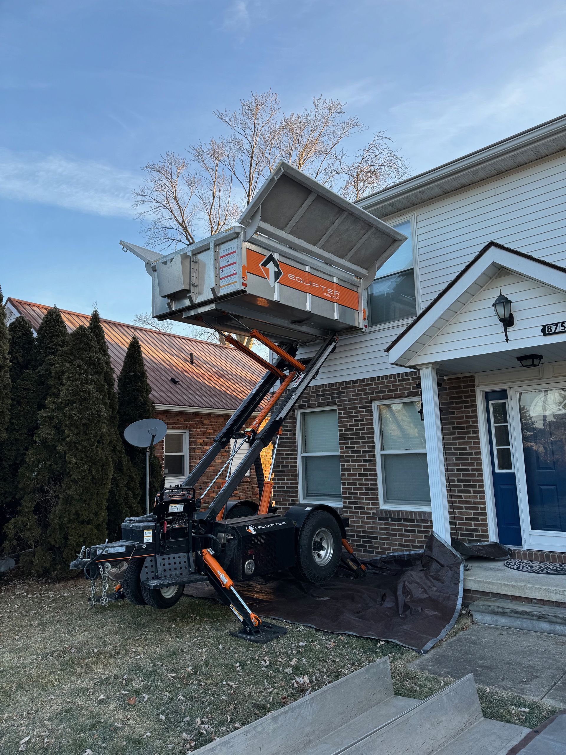 A truck-mounted lift is raised to a house for moving items. An orange container is in position to receive the items.