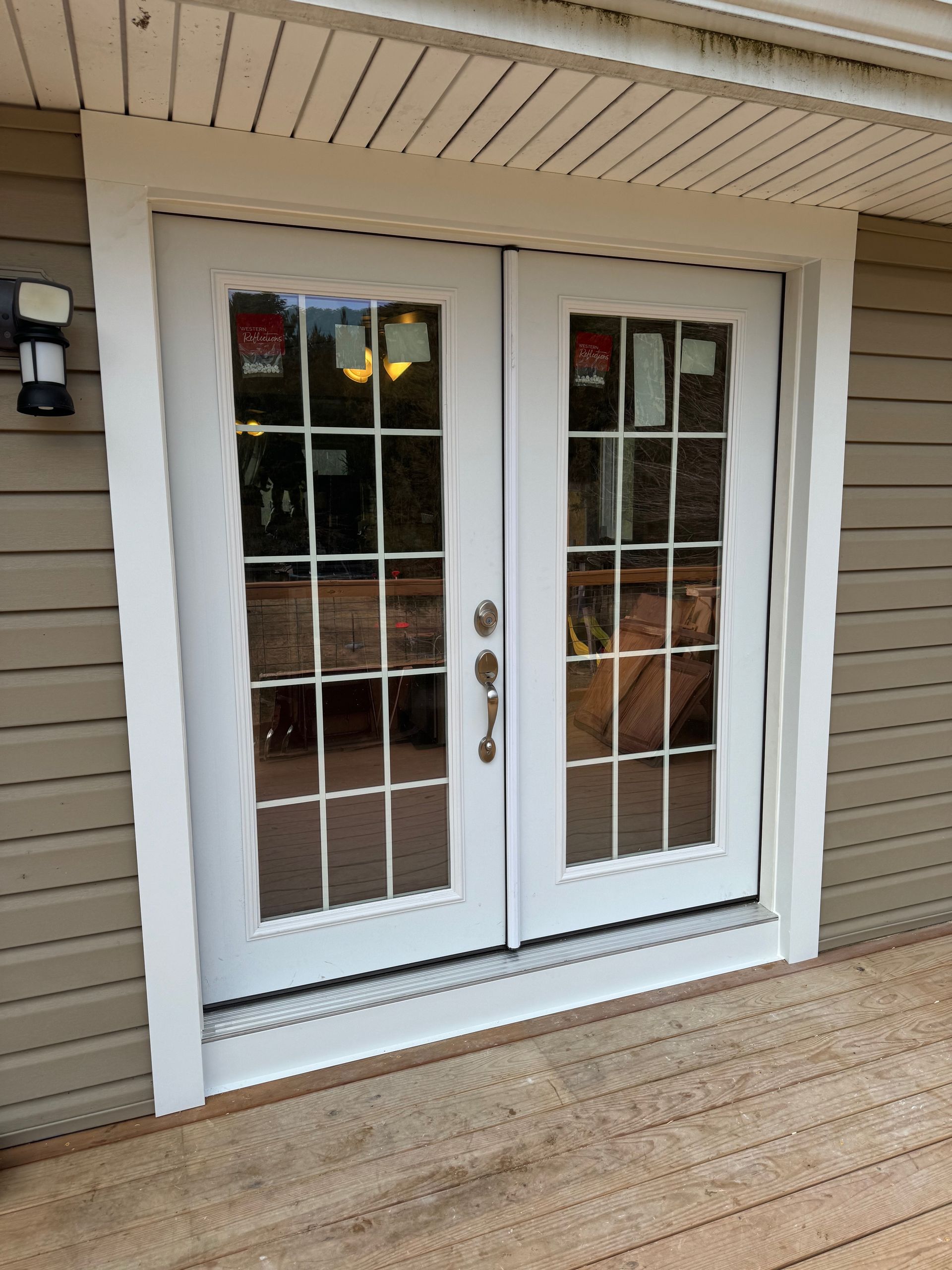 White double French doors with grid windows, trimmed in white, set into beige siding, on a wooden deck.