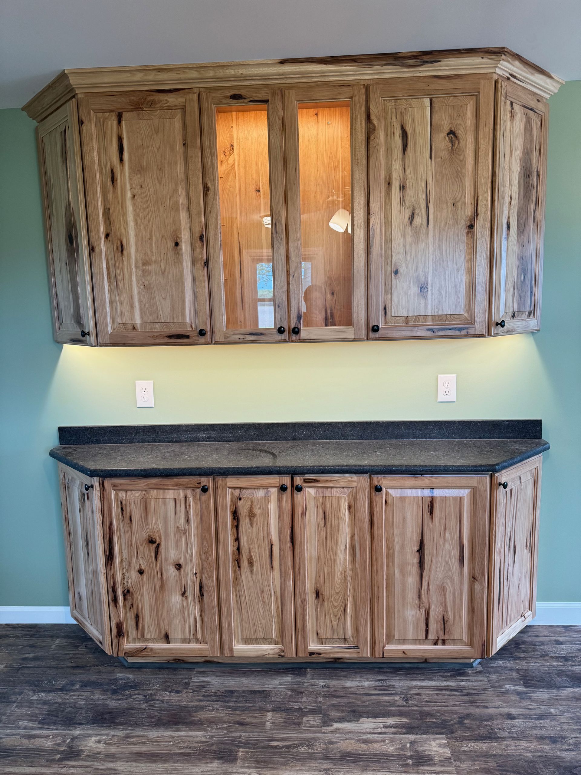 Rustic wooden cabinets with dark countertop, mounted on green wall with under-cabinet lighting.