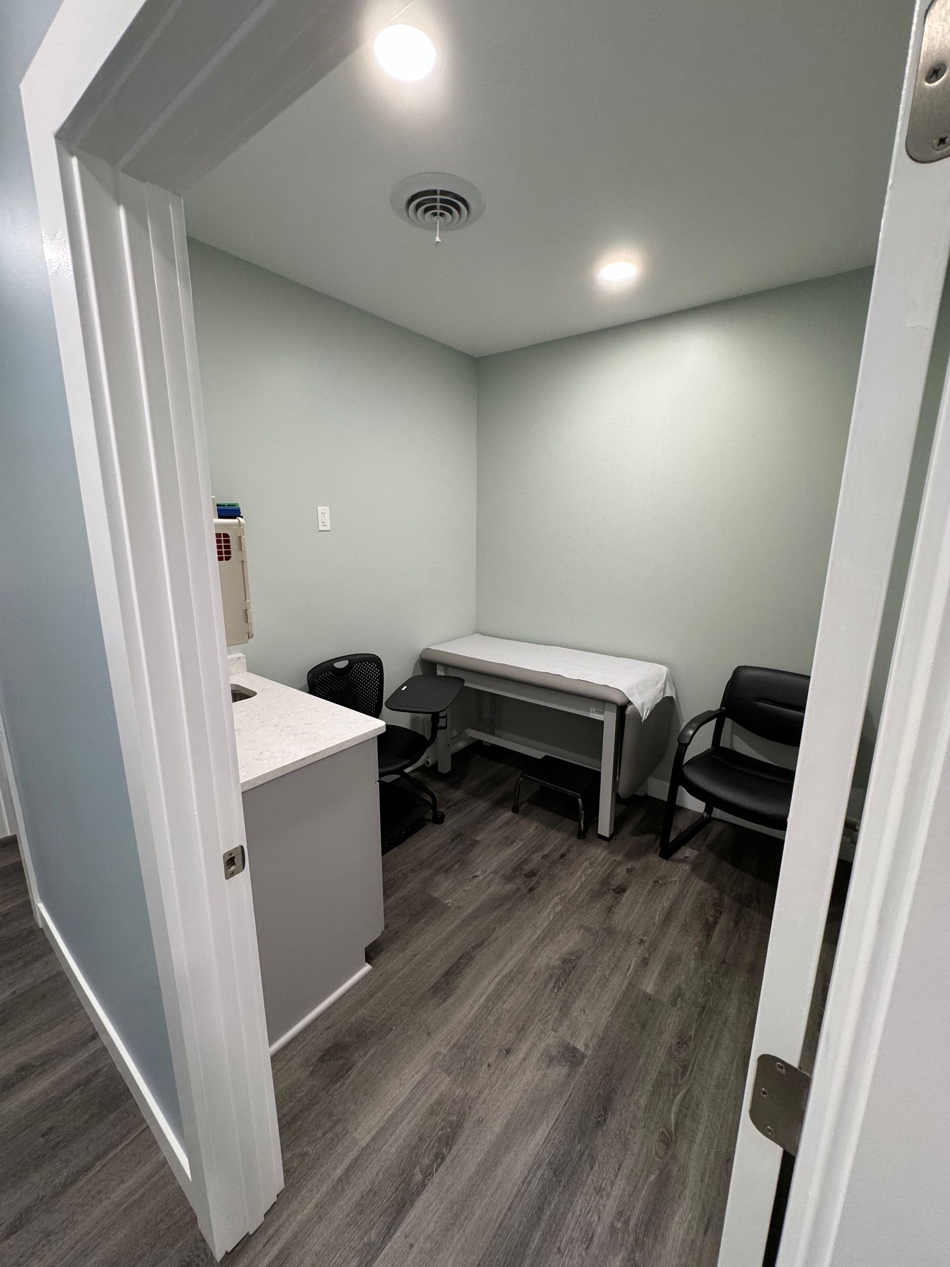 Medical exam room with examination table, sink, and chair; grey and light blue.