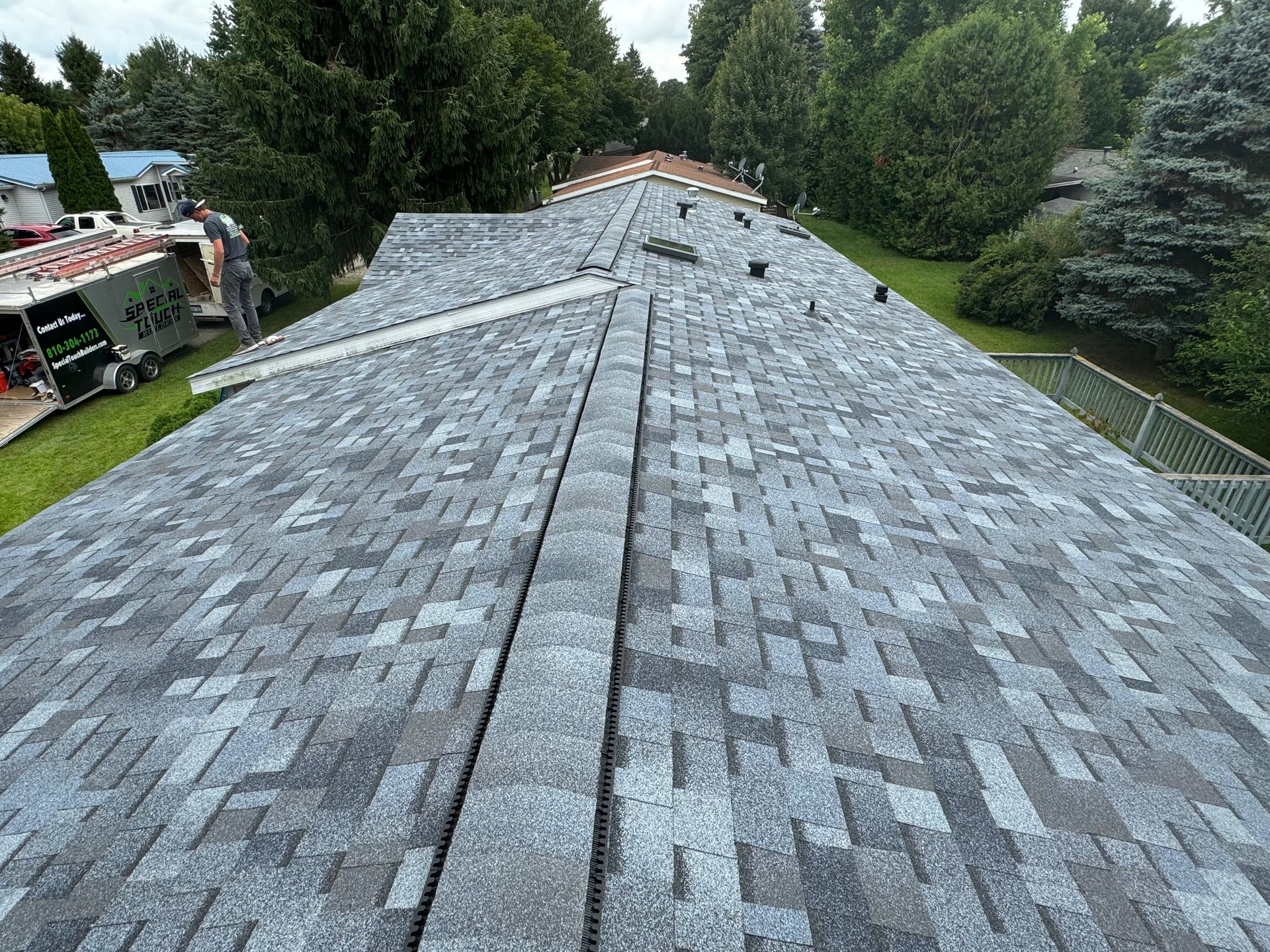 Rooftop with gray shingles; a person stands near a truck, working. Green grass and trees in the background.