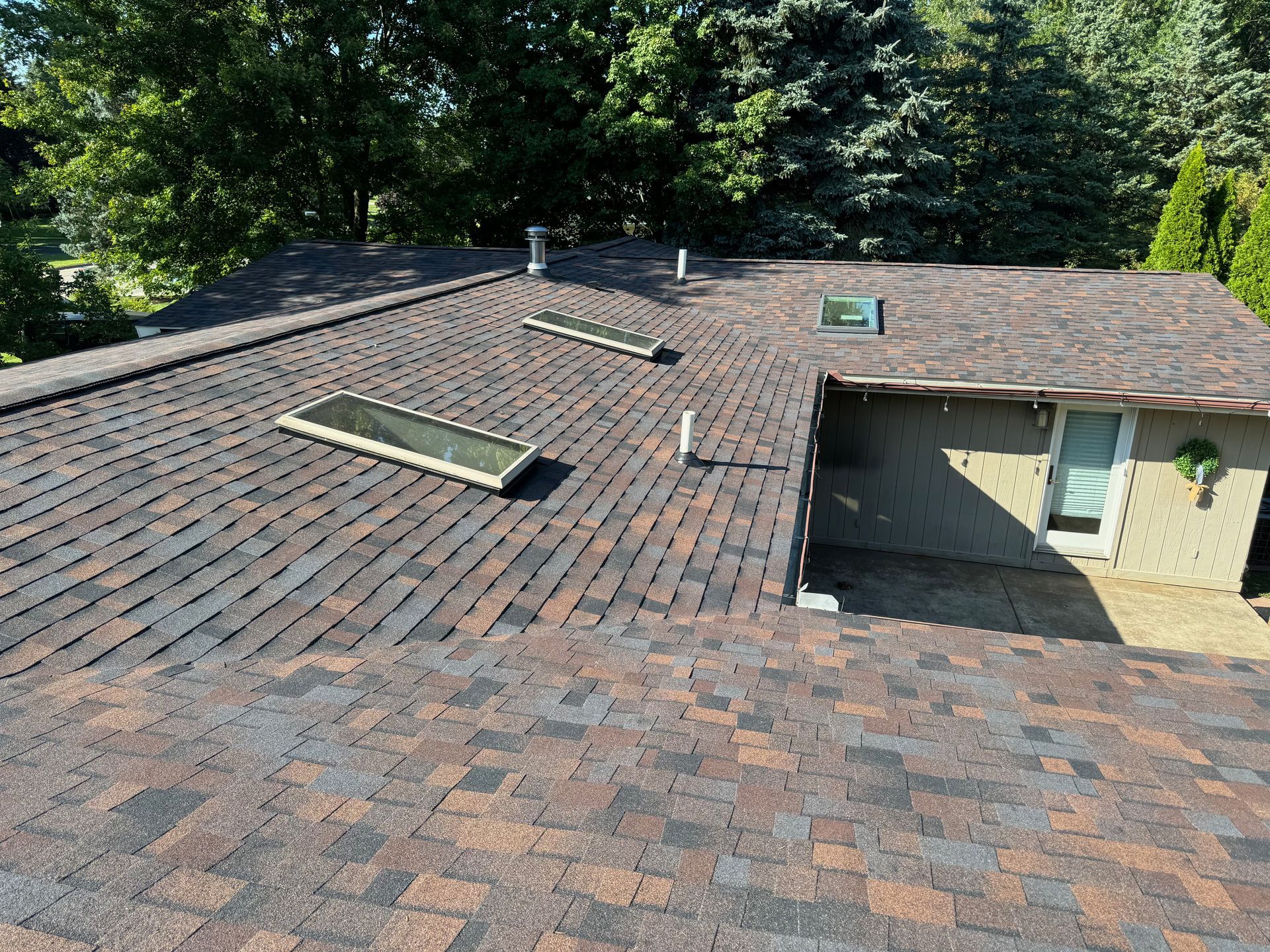 Brown asphalt shingle roof with skylights and vents. Overlooks a small building and trees.