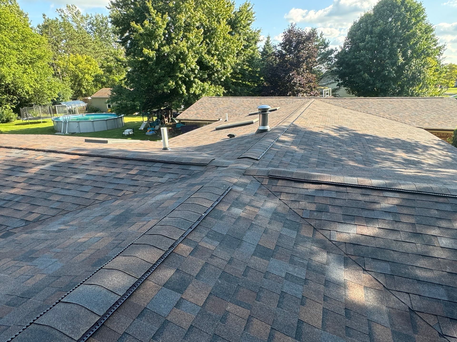 View of a brown, multi-toned asphalt shingle roof on a sunny day with trees in the background.