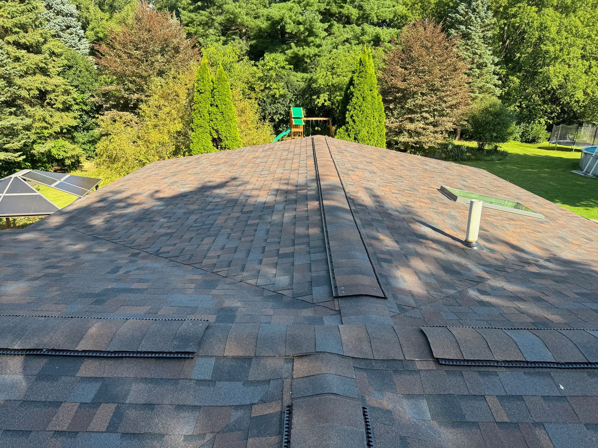 View of a brown asphalt shingle roof with a central ridge, trees in the background.