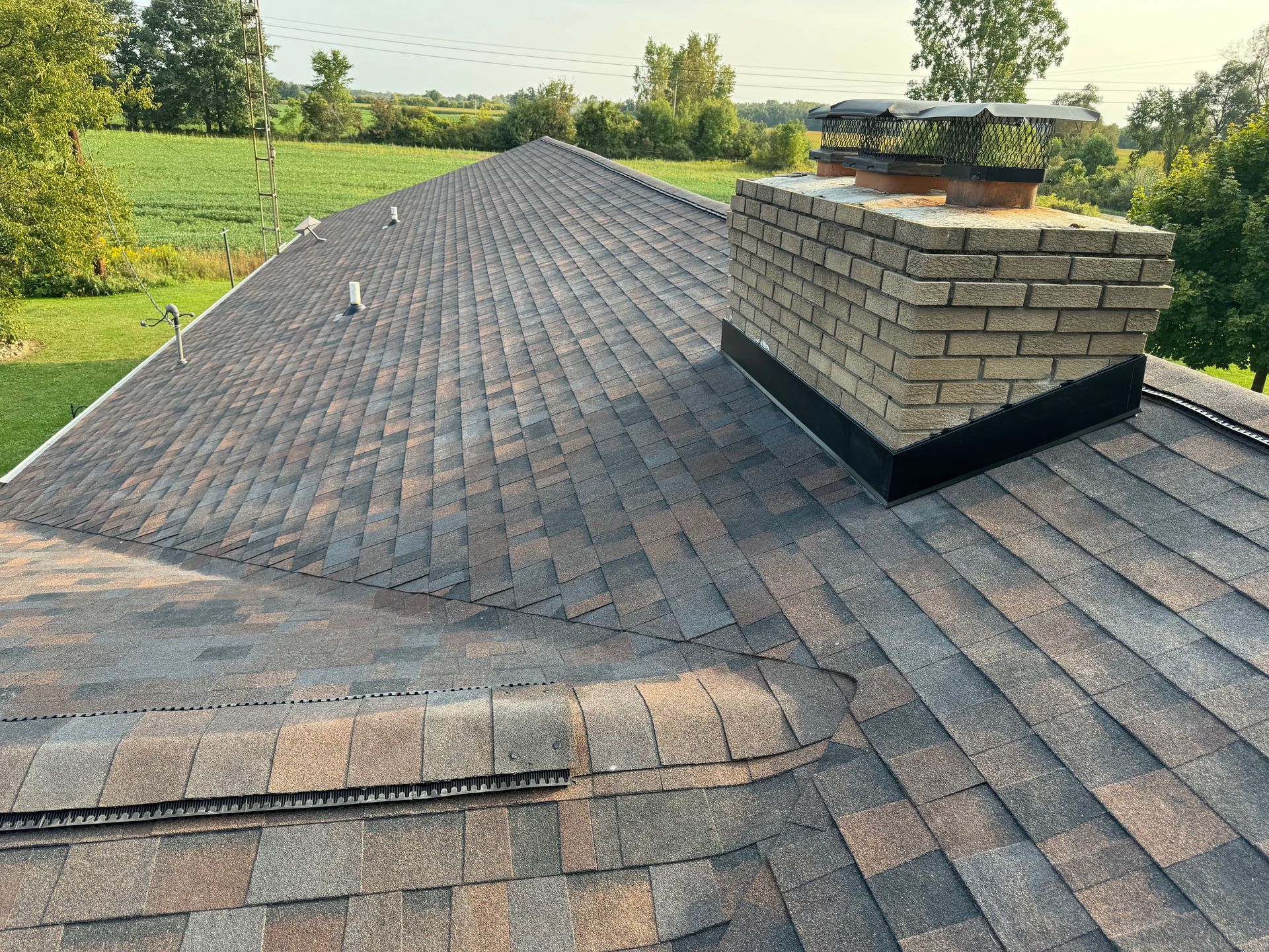 A roof with asphalt shingles, a brick chimney, and a rural landscape in the background.