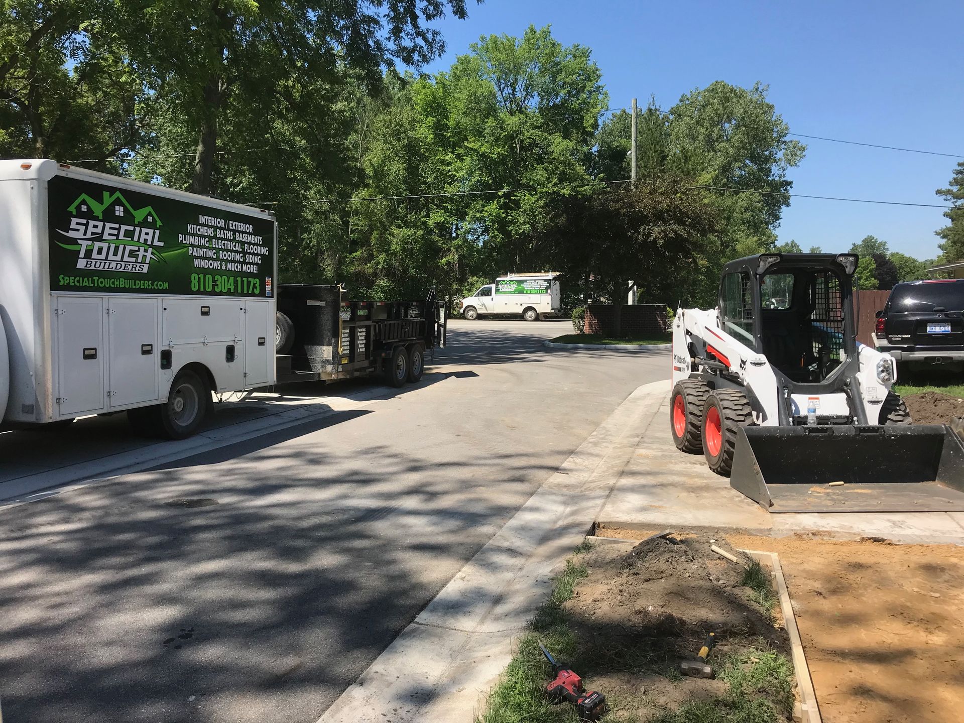 A landscape company trailer and Bobcat in a driveway with trees and a sidewalk.