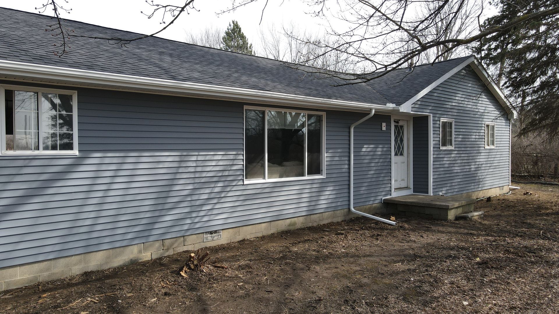 Blue-sided house with white trim, windows, and door, surrounded by bare ground and trees.