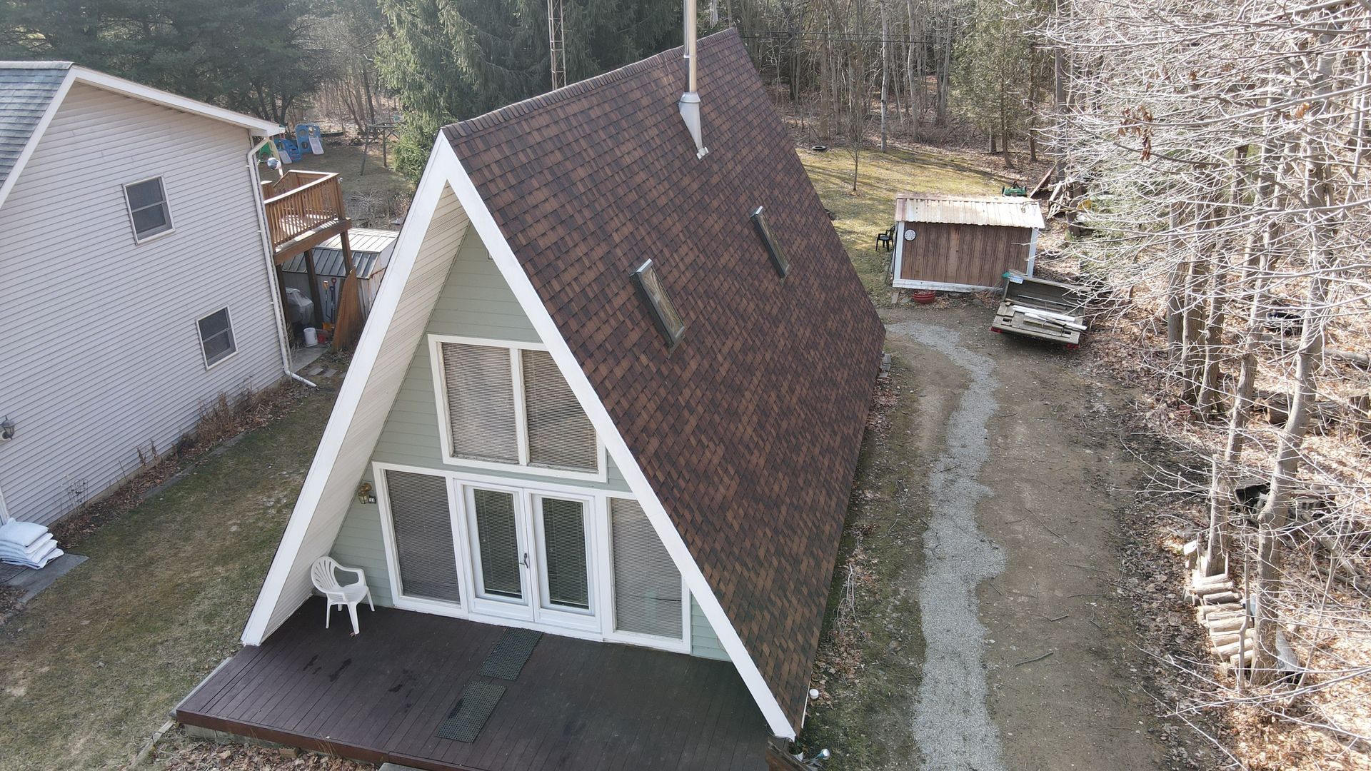 A-frame house with brown roof and light green siding, white trim, and a car parked on the gravel driveway.