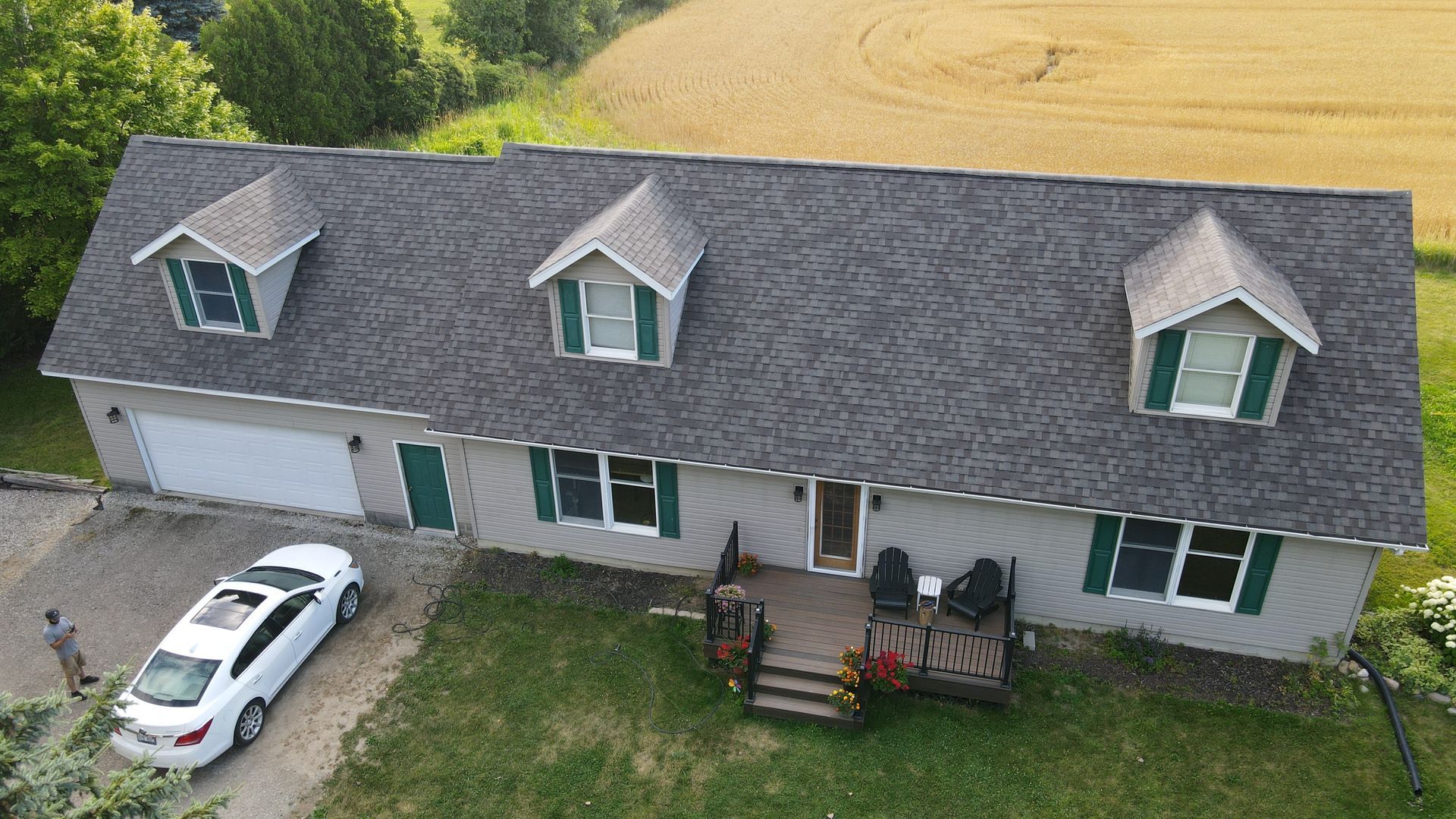 Gray house with three dormers, garage, white car, person, and grassy yard.