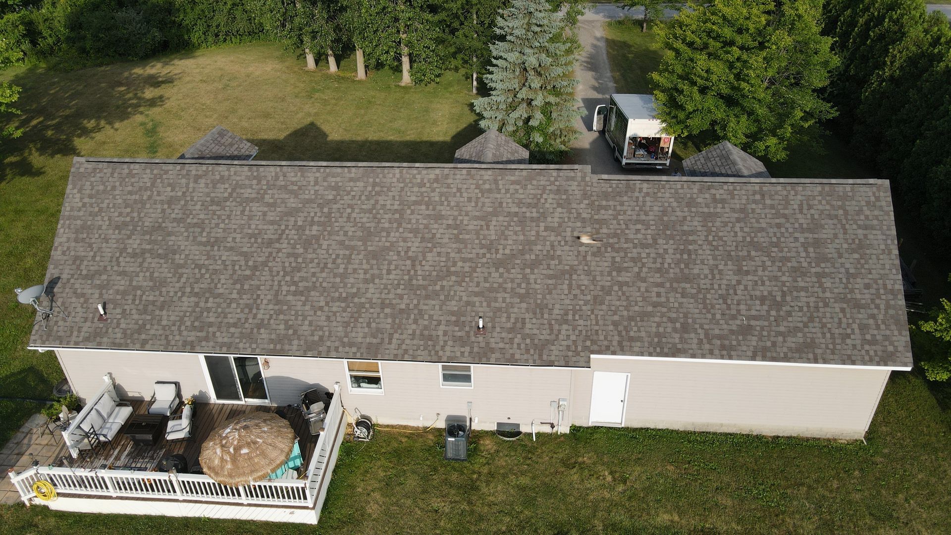 Overhead view of a house with a gray roof, deck, and a truck parked nearby. Green yard and trees surround.