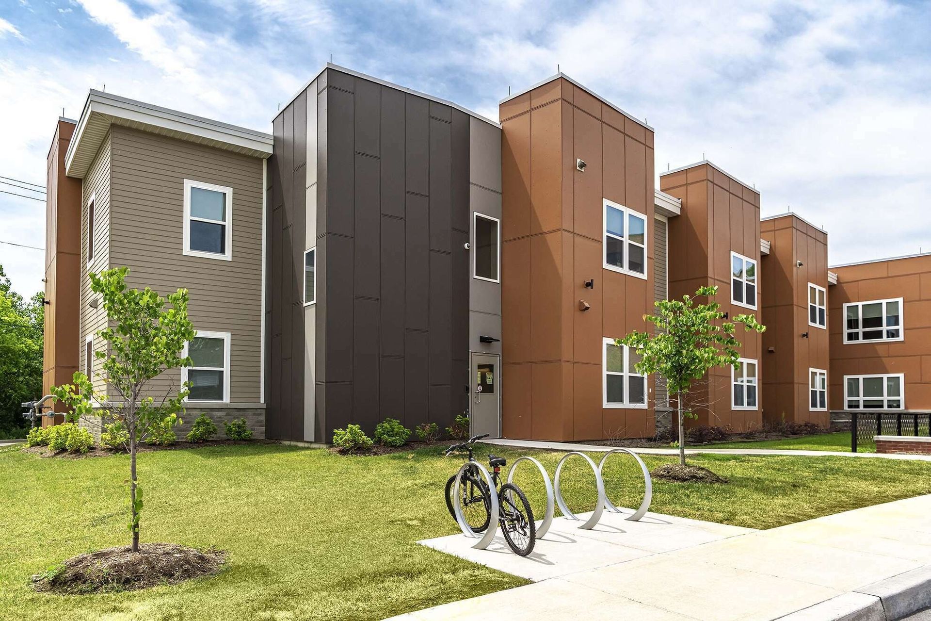 Modern apartment building with brown and gray facades, grass lawn, sidewalk, and bike rack in front.