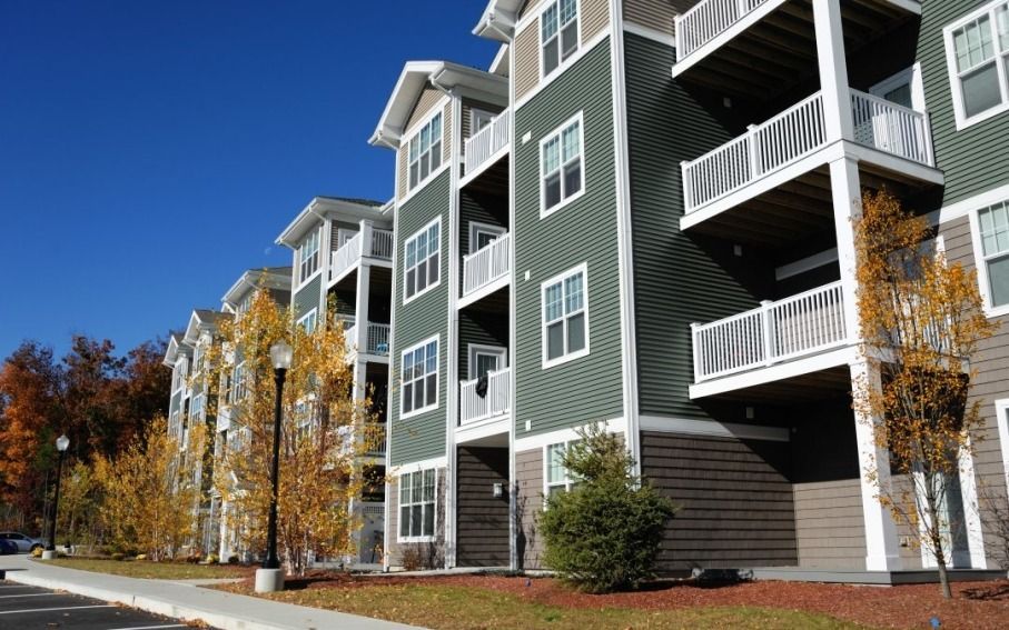 Apartment buildings with green siding and white balconies beside a tree-lined walkway on a clear day