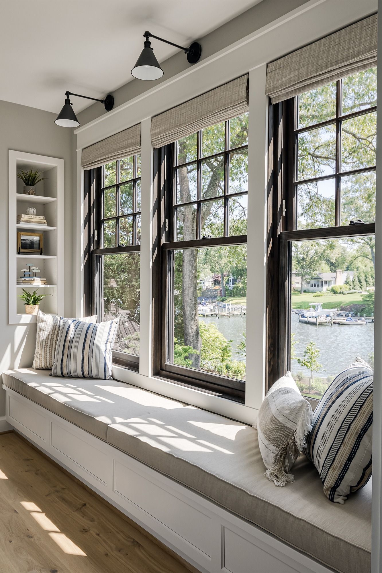 Bright sunlit window seat with cushions, black-framed windows, and a view of trees and water outside