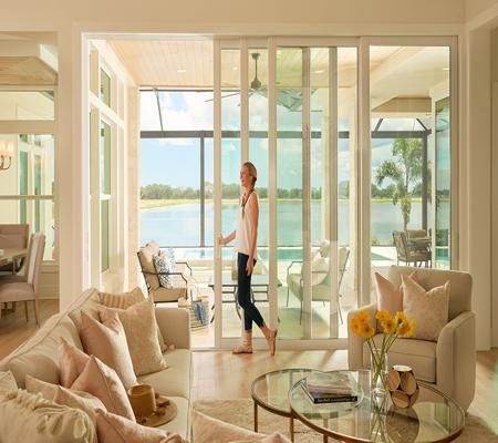 Bright living room with glass doors opening to a sunlit waterfront patio; one person walking through.