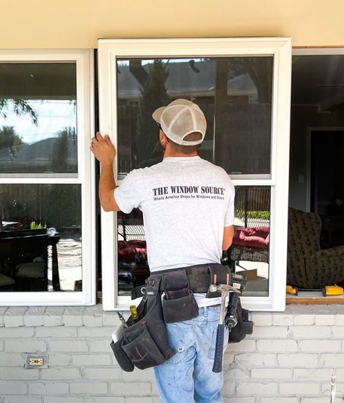 Worker installing a window on a house exterior, wearing a white T-shirt and tool belt.