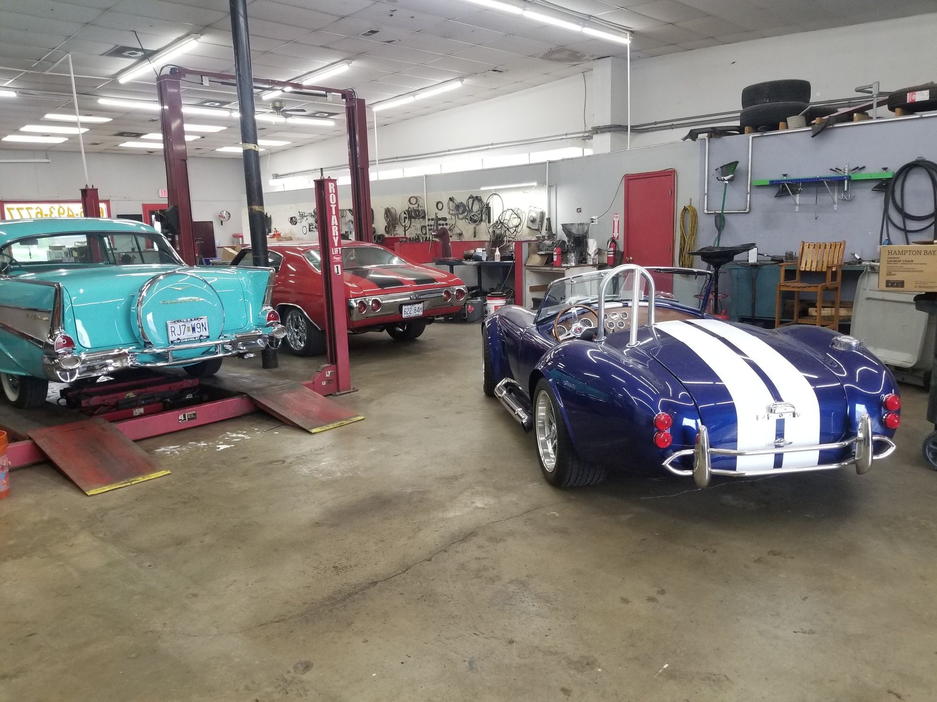 Cars in a garage: a blue and white Cobra, a blue classic car on a lift, and a red car in the background.