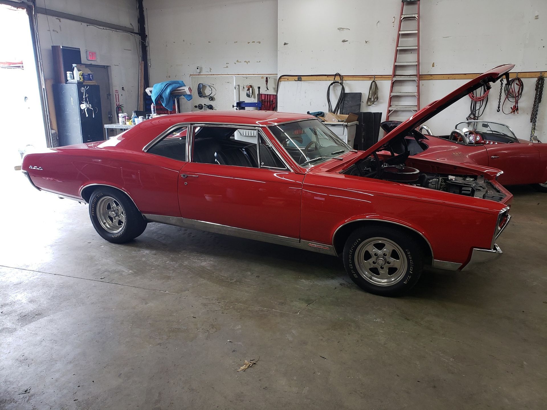 Red 1967 Pontiac GTO coupe with open hood in a garage, chrome trim, silver wheels.