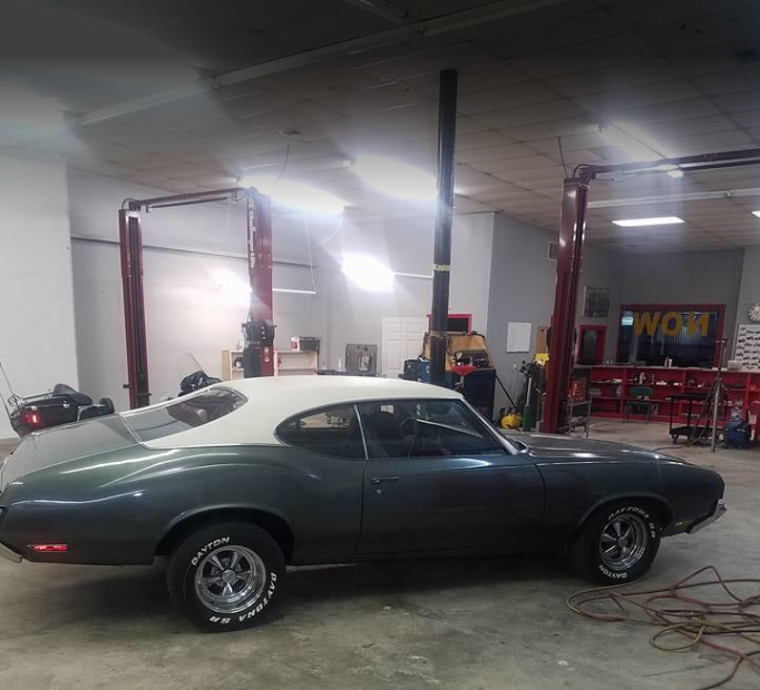 A classic dark green Oldsmobile Cutlass coupe with a white roof in a garage with a car lift.