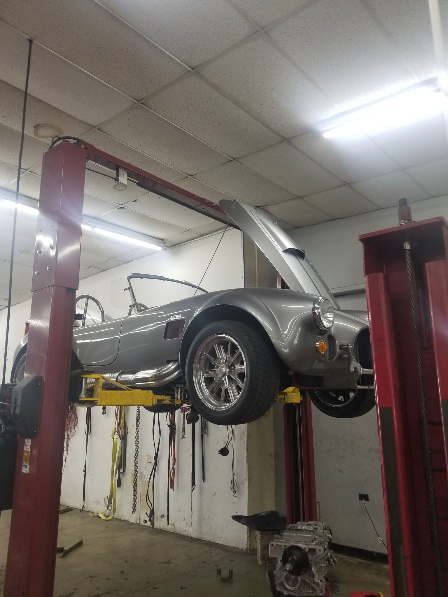 Silver sports car raised on a lift in a garage, hood open for repair.