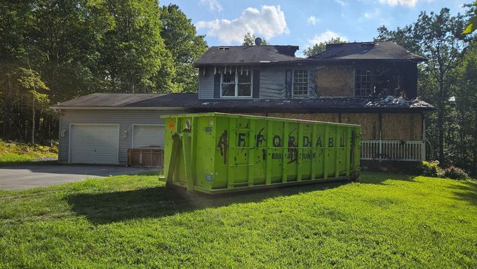 Two-story house with boarded-up windows, a green dumpster in the yard, and a garage.