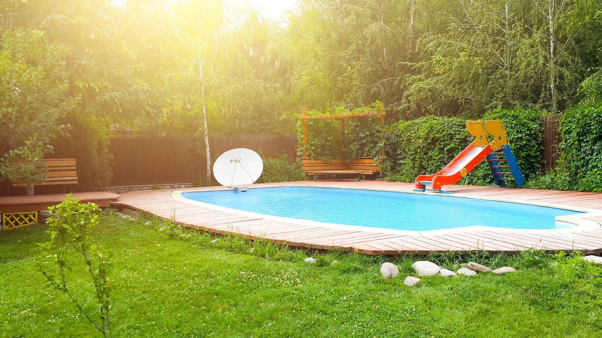 Sunny backyard pool with blue water, orange slide, white lounge chair, and lush green lawn and trees