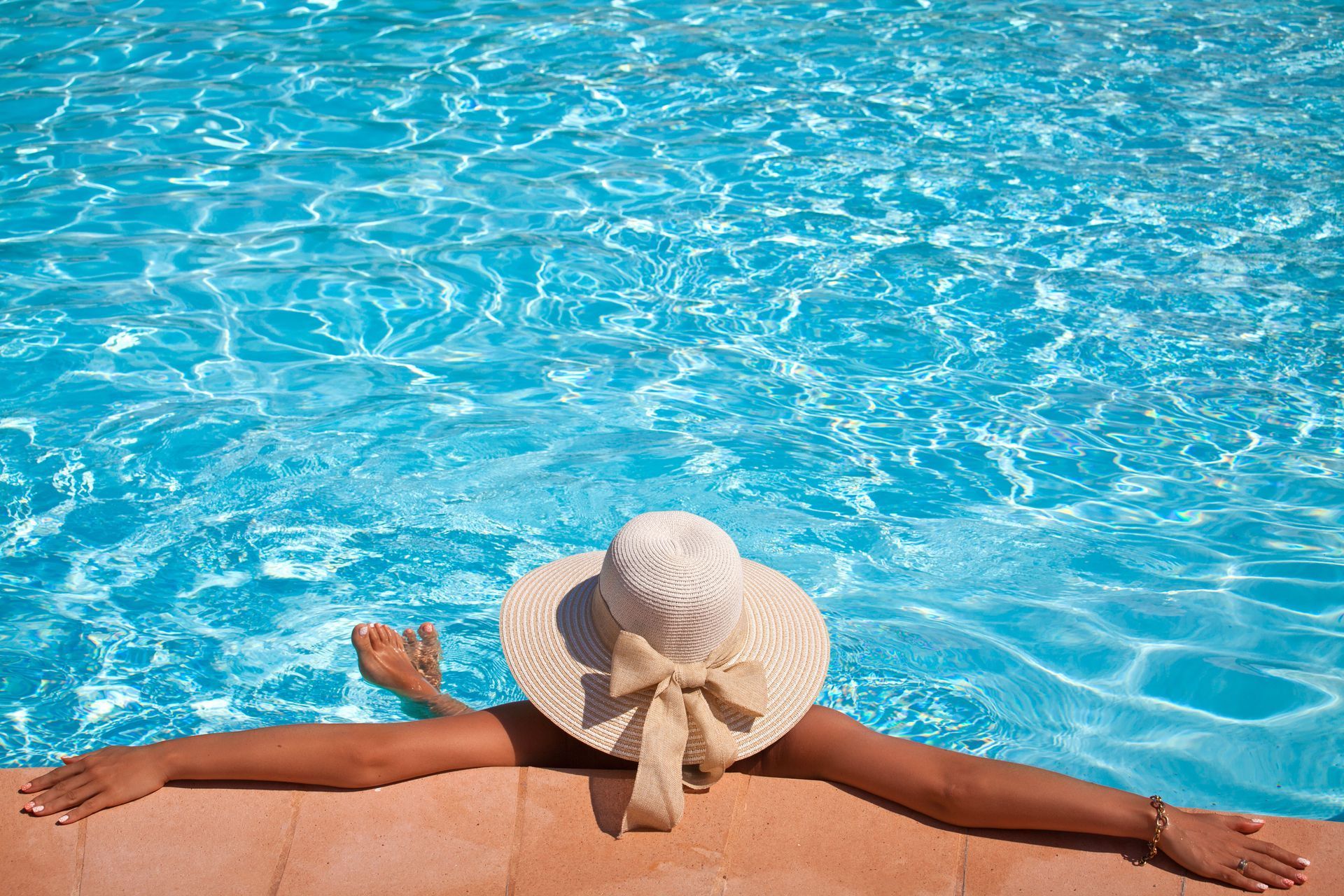 Person relaxing at the edge of a bright blue swimming pool, wearing a sunhat and stretching out arms