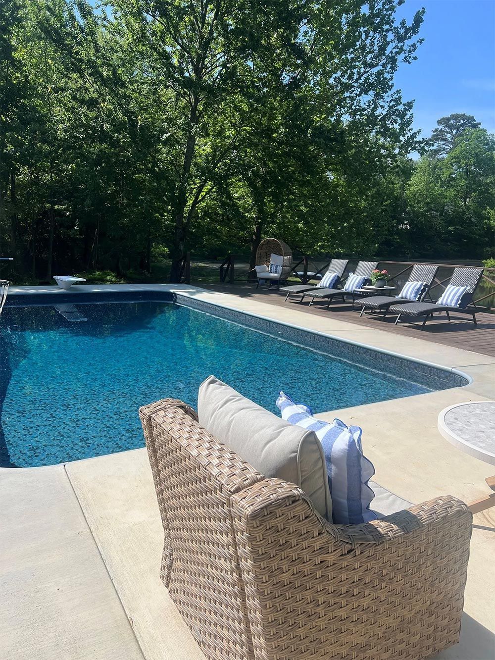 Poolside patio with blue swimming pool, wicker chair, lounge chairs, and trees in bright sunlight