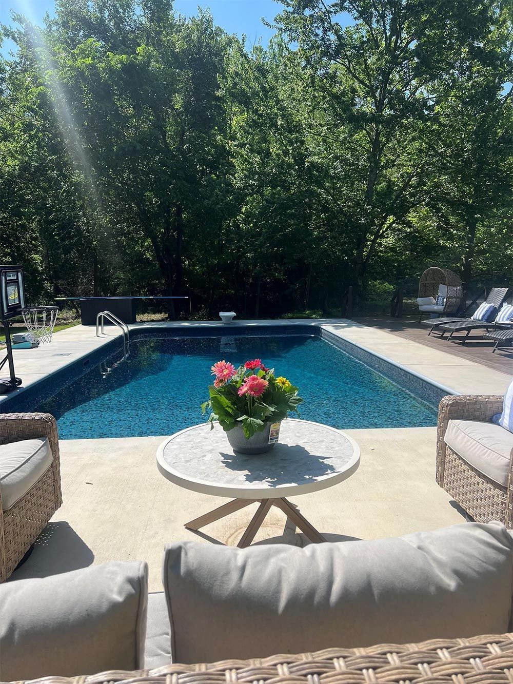 Backyard pool with patio seating, a flower arrangement on a table, and trees in bright sunlight