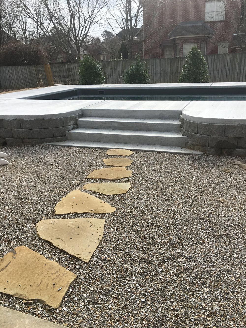 Stepping-stone path leads across gravel to gray stone steps and a raised patio in a backyard.