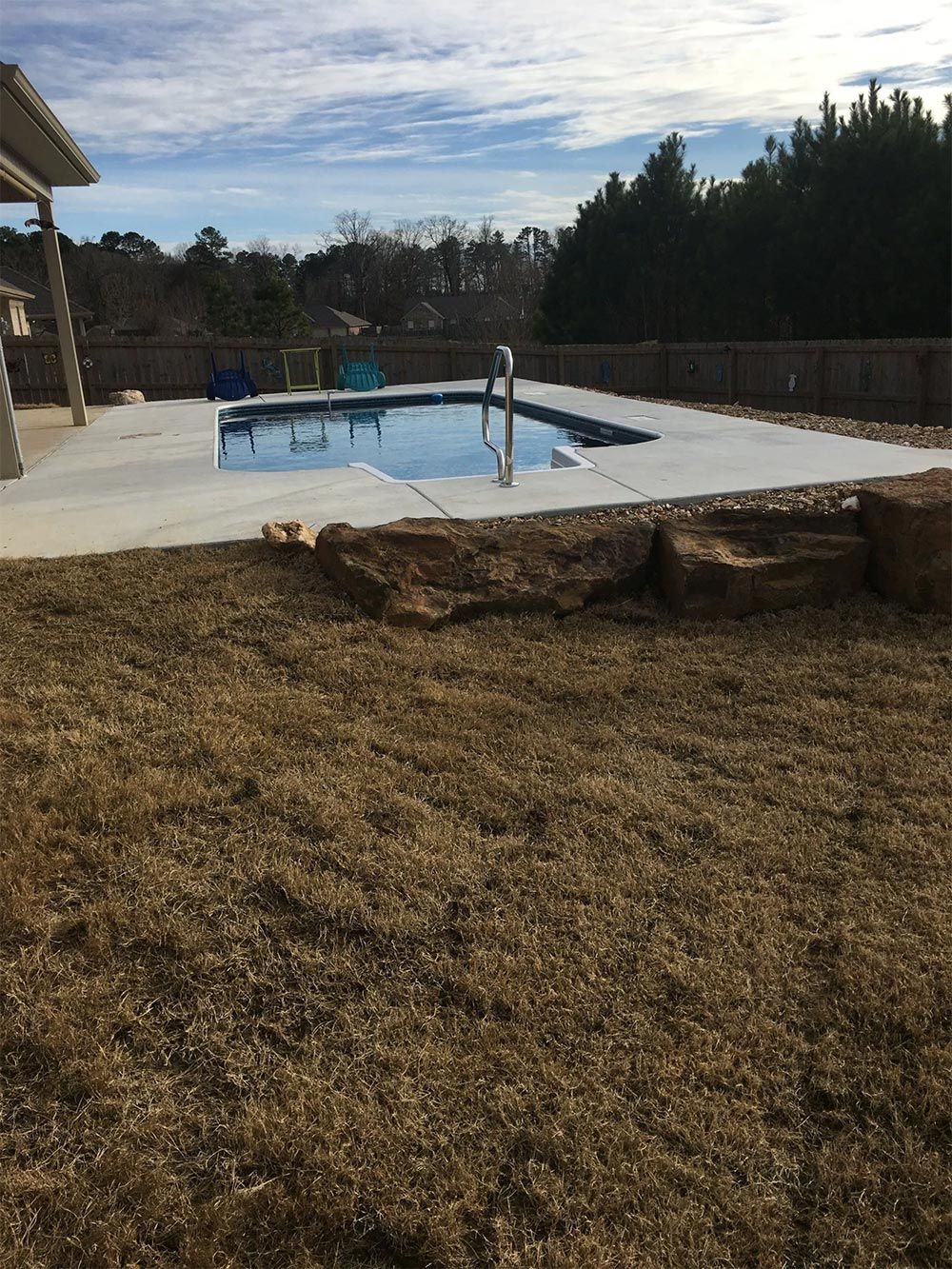 Above-ground pool on a concrete deck in a backyard, surrounded by grass and a wooden fence.