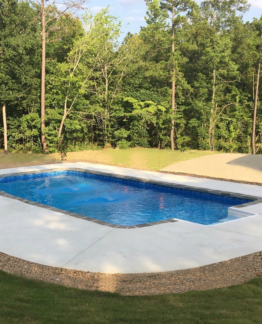 Backyard swimming pool with blue water, concrete deck, and trees in the background
