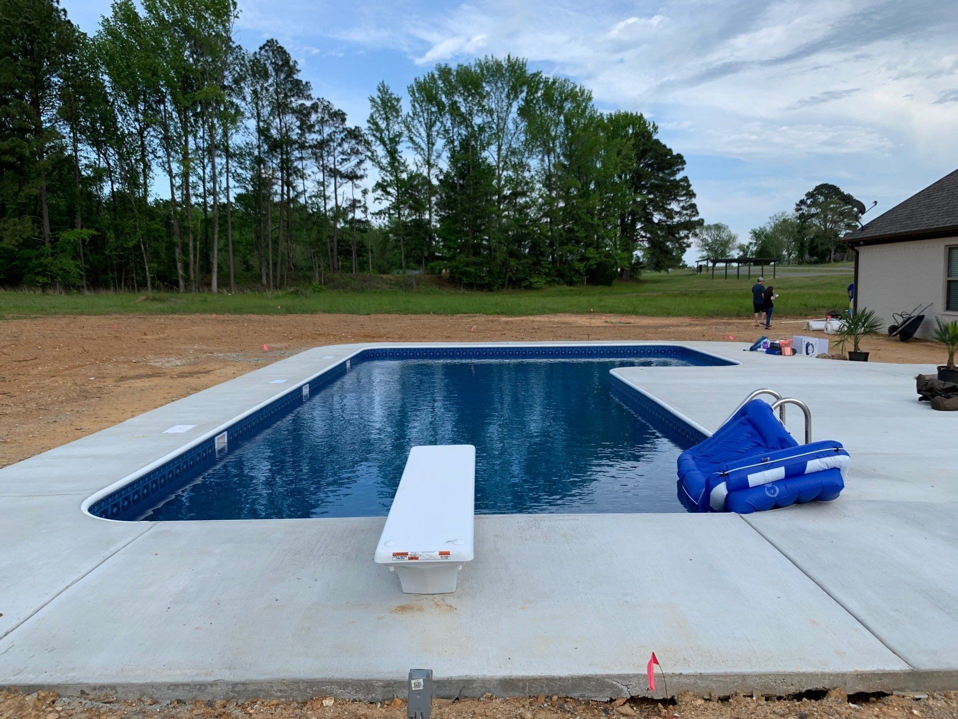 Rectangular backyard swimming pool with a diving board and blue pool float on the deck, trees in the background