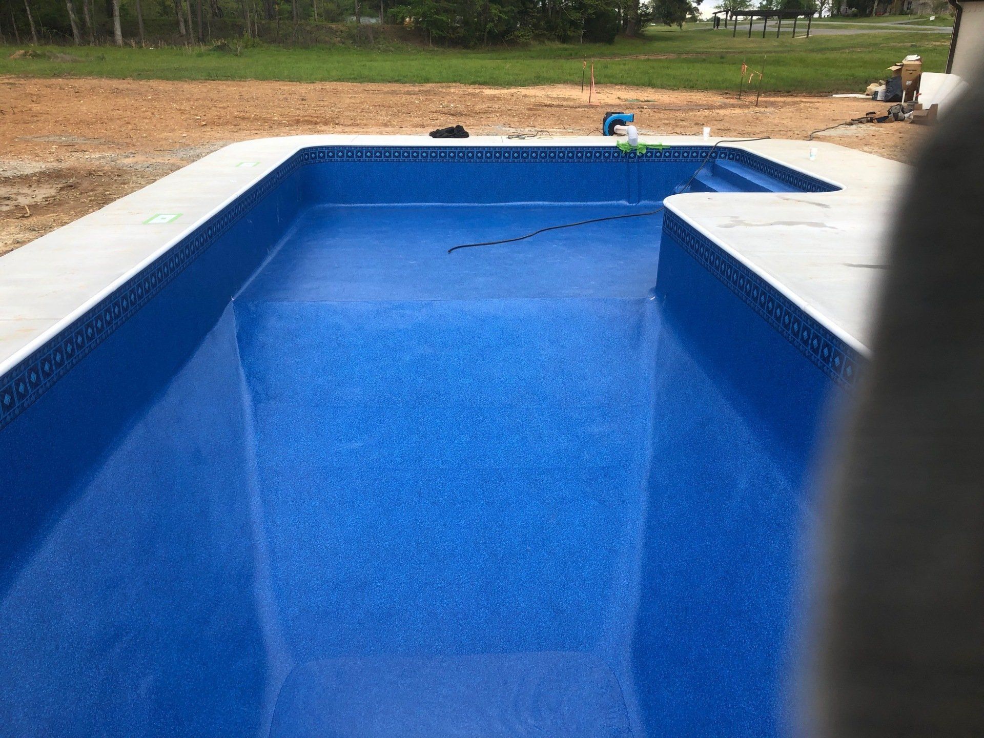 Empty blue swimming pool under construction with surrounding concrete deck and grassy yard background