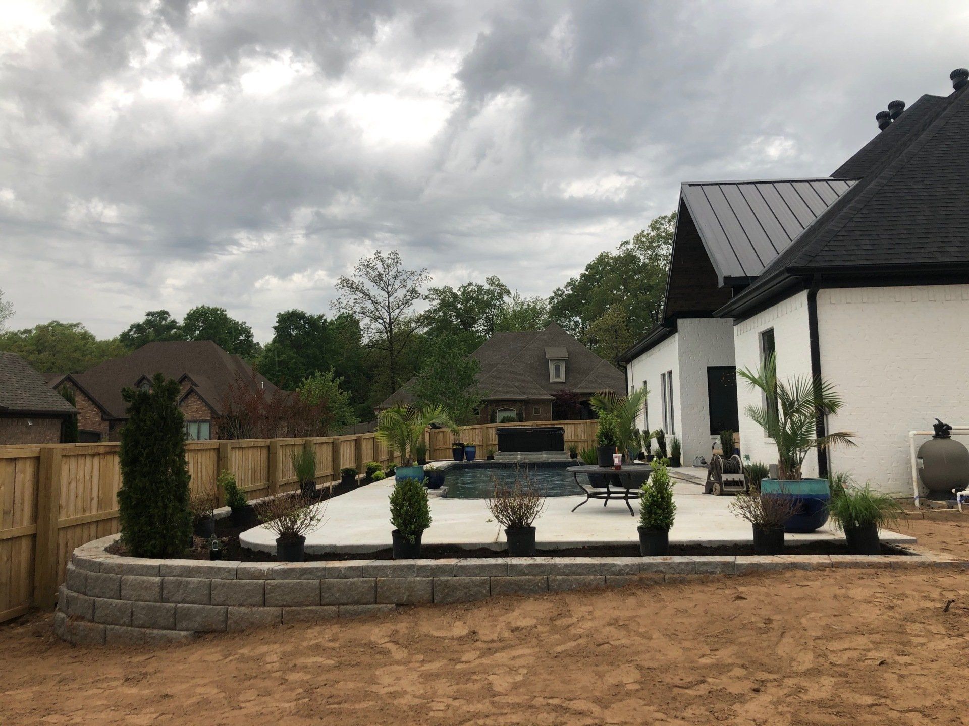 Backyard patio with raised circular planter, potted shrubs, and a white house under cloudy skies