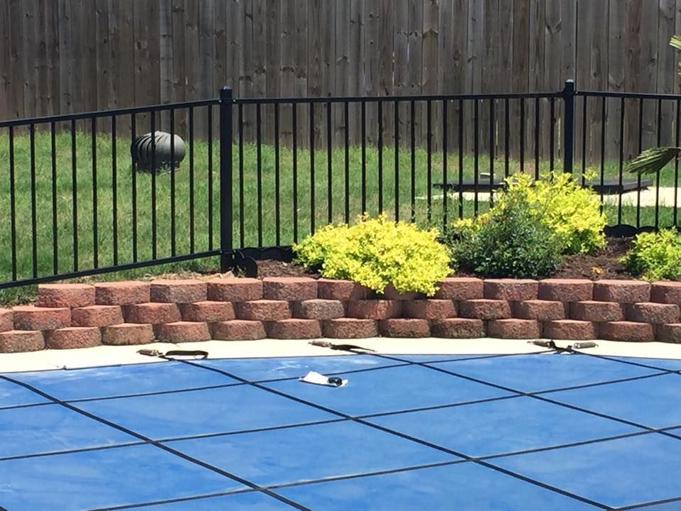 Backyard pool with blue cover, brick retaining wall, yellow shrubs, and a wooden fence
