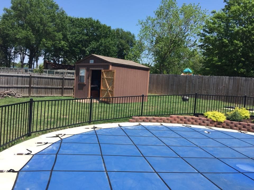Backyard with a covered blue pool, brown shed, black fence, lawn, and trees under a sunny sky