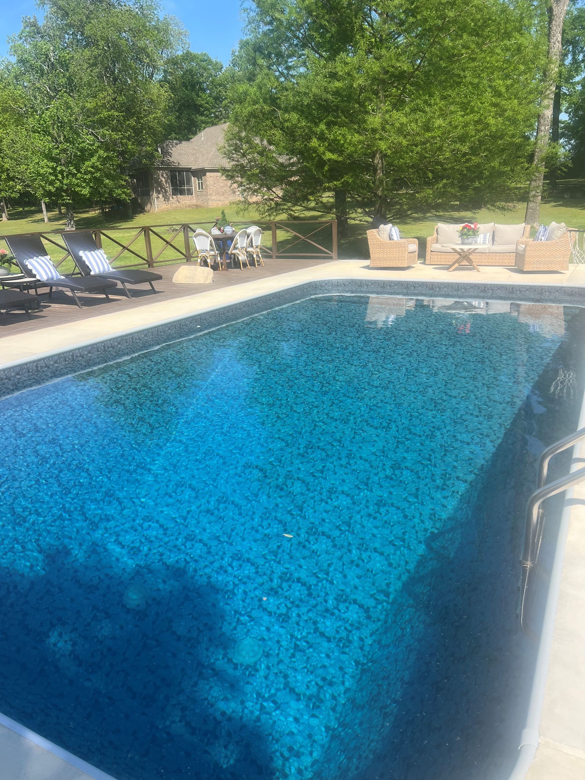 Backyard swimming pool with lounge chairs, trees, and a stone patio in bright sunlight