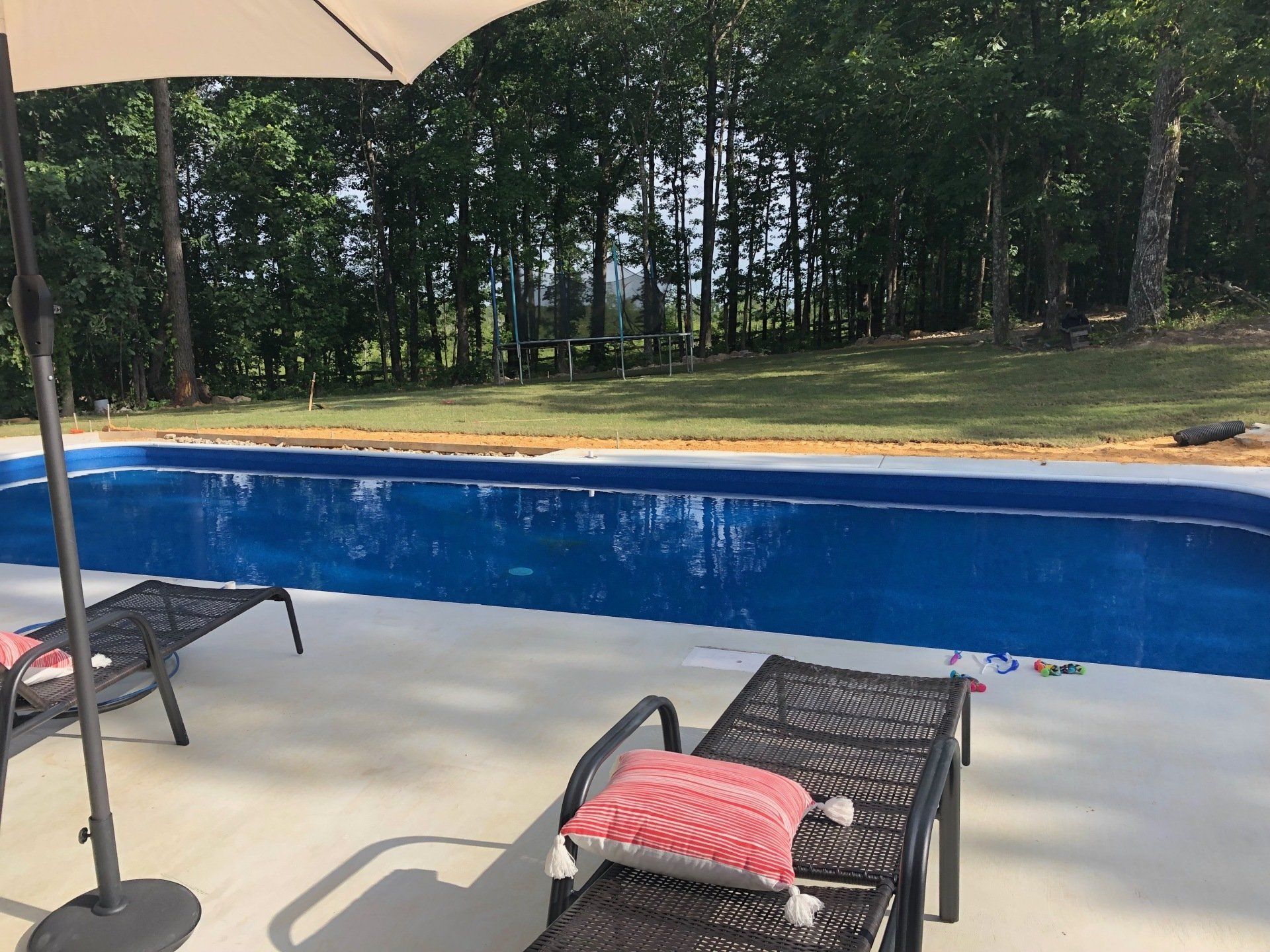 Patio lounge chairs beside a blue pool with an umbrella and wooded backyard backdrop
