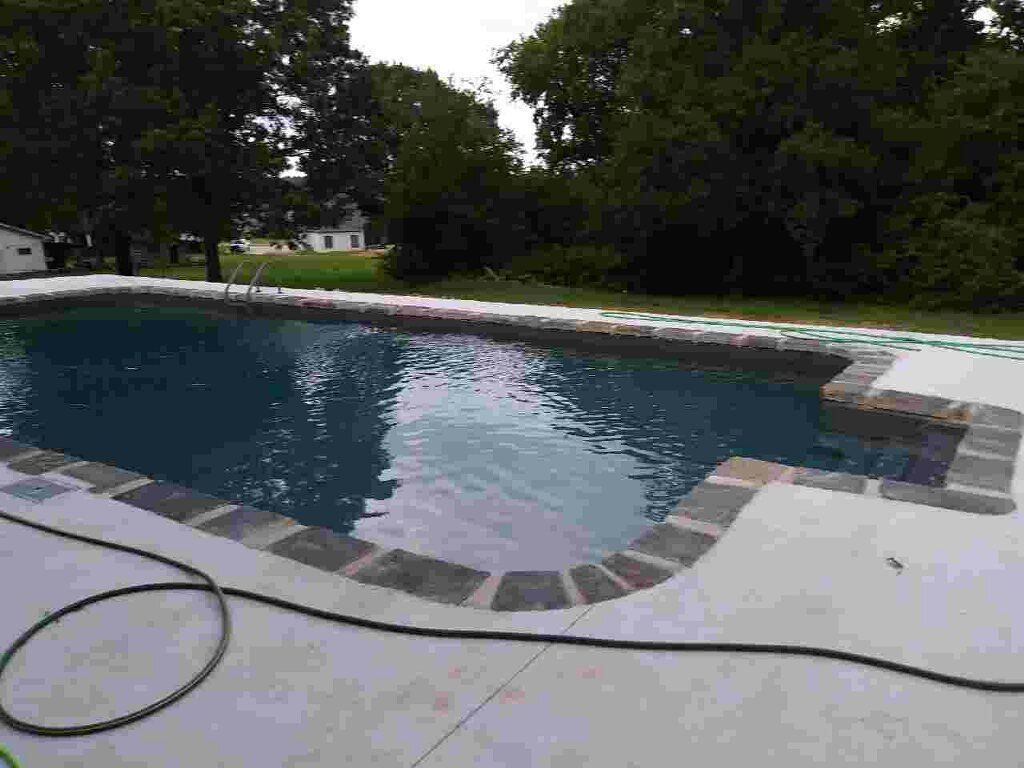 Backyard swimming pool with curved stone edge, surrounded by trees and concrete deck