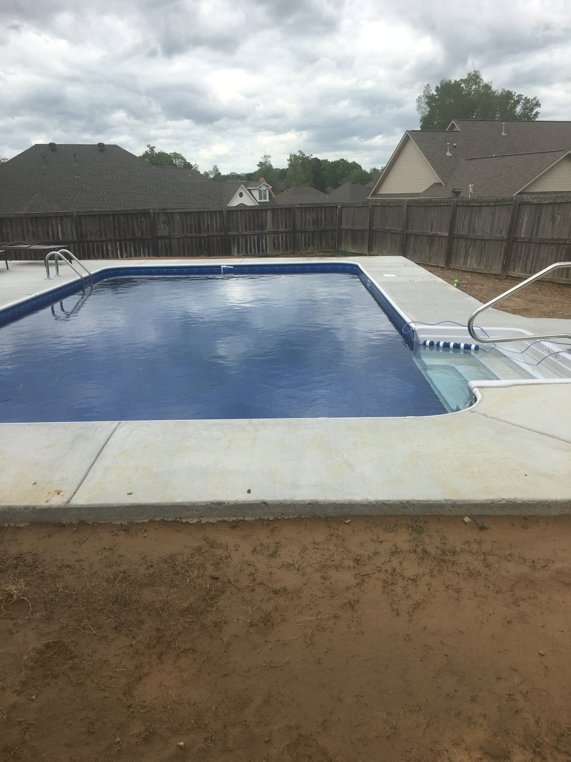 Rectangular backyard swimming pool with blue water, concrete deck, and wooden fence under a cloudy sky
