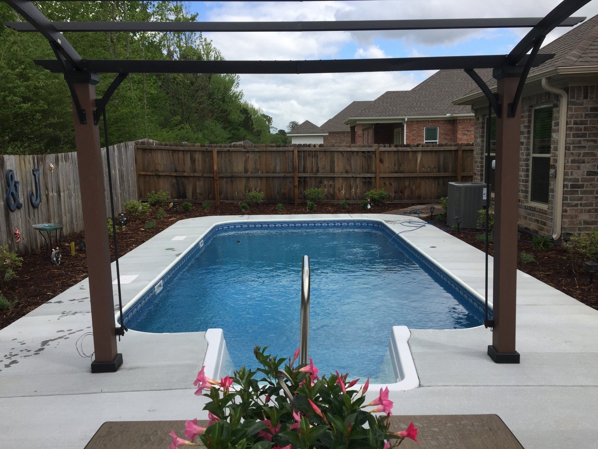 Backyard swimming pool with white deck, pergola, wooden fence, and pink flowers in the foreground