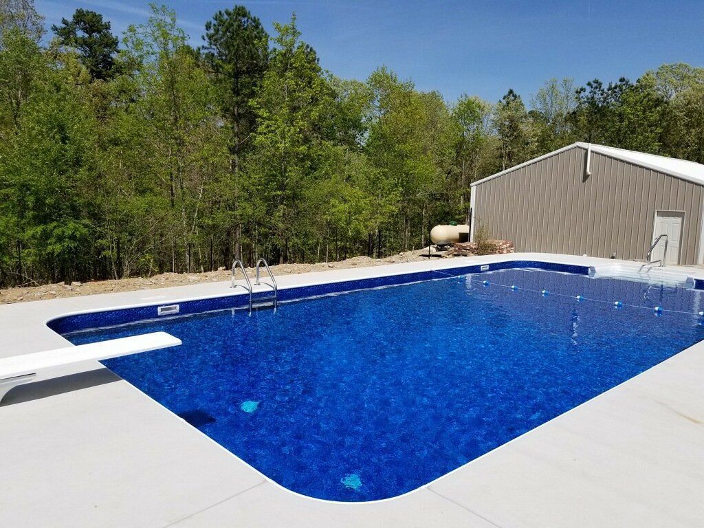 Outdoor backyard swimming pool with blue water, white deck, and surrounding trees under a clear sky