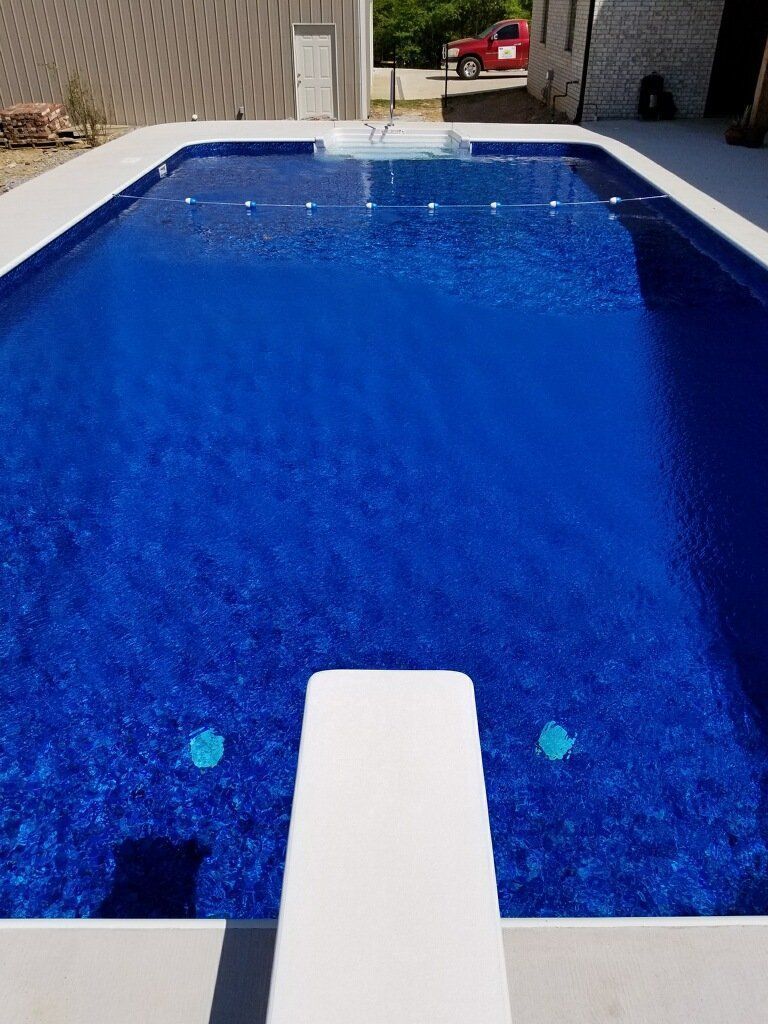 Blue swimming pool with a diving board in the foreground and a red car in the background