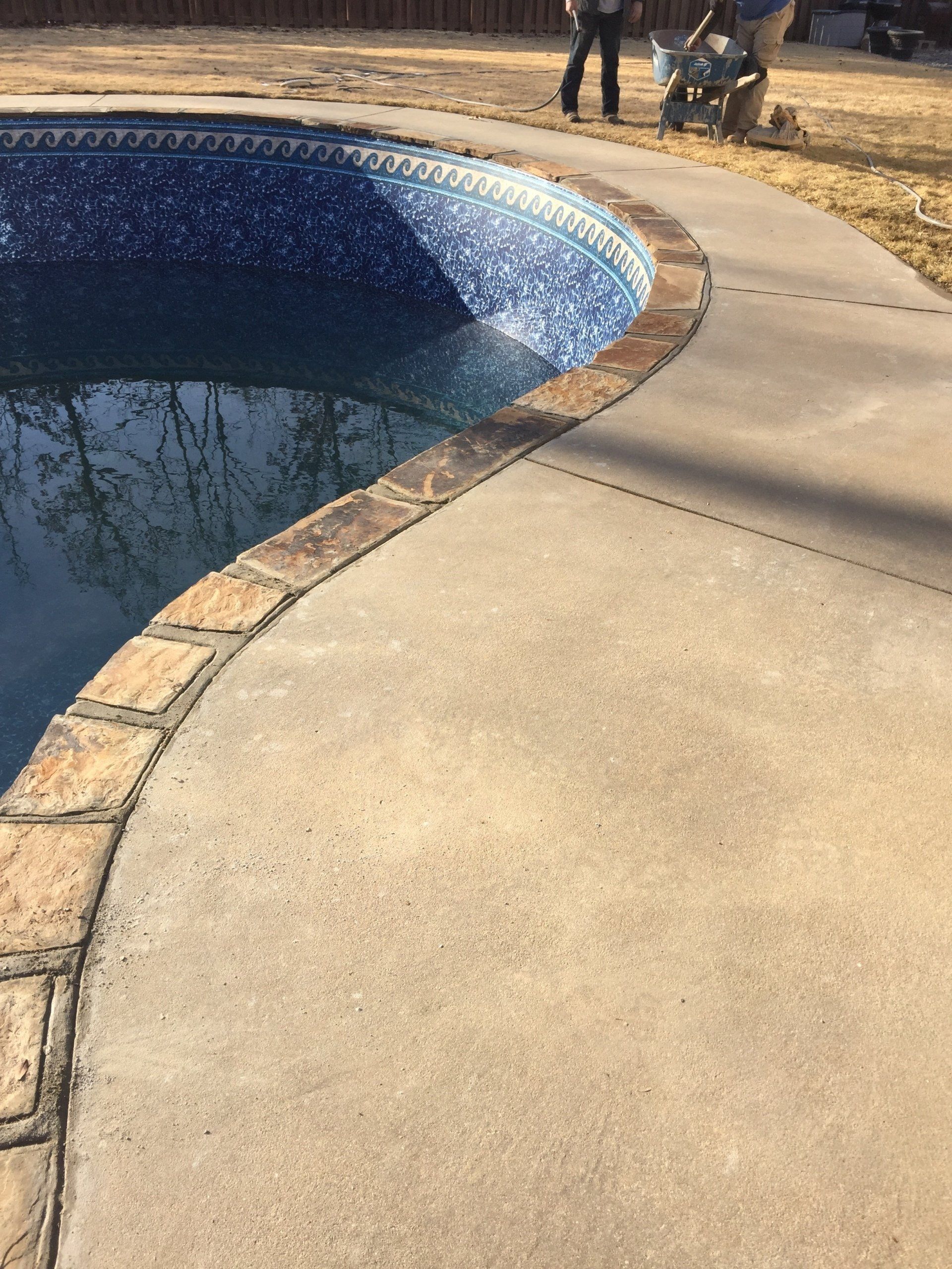 Curved concrete pool deck beside a blue-tiled swimming pool, with a few people in the background.