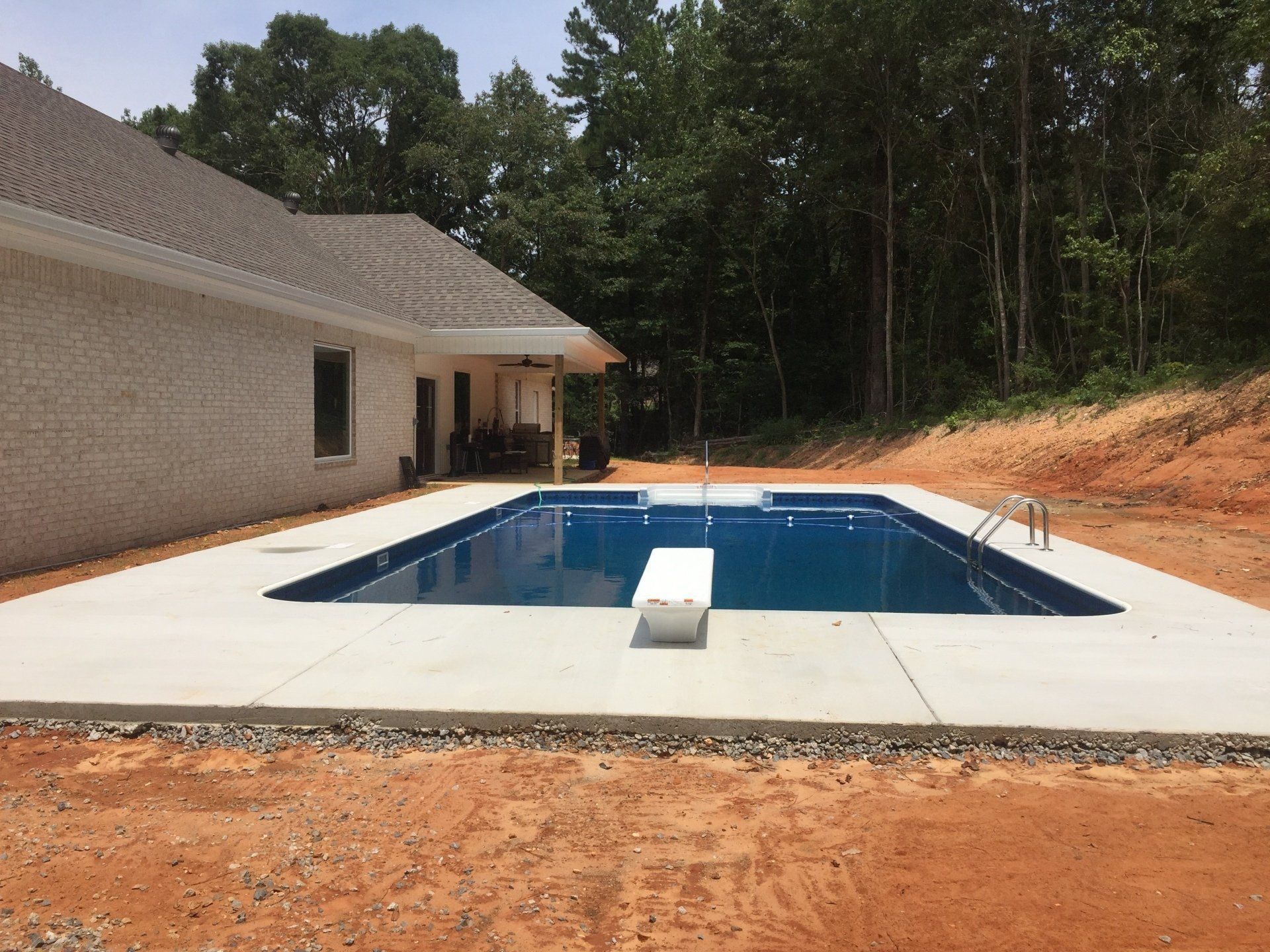 Backyard with a rectangular in-ground pool beside a house, bordered by concrete and red dirt.