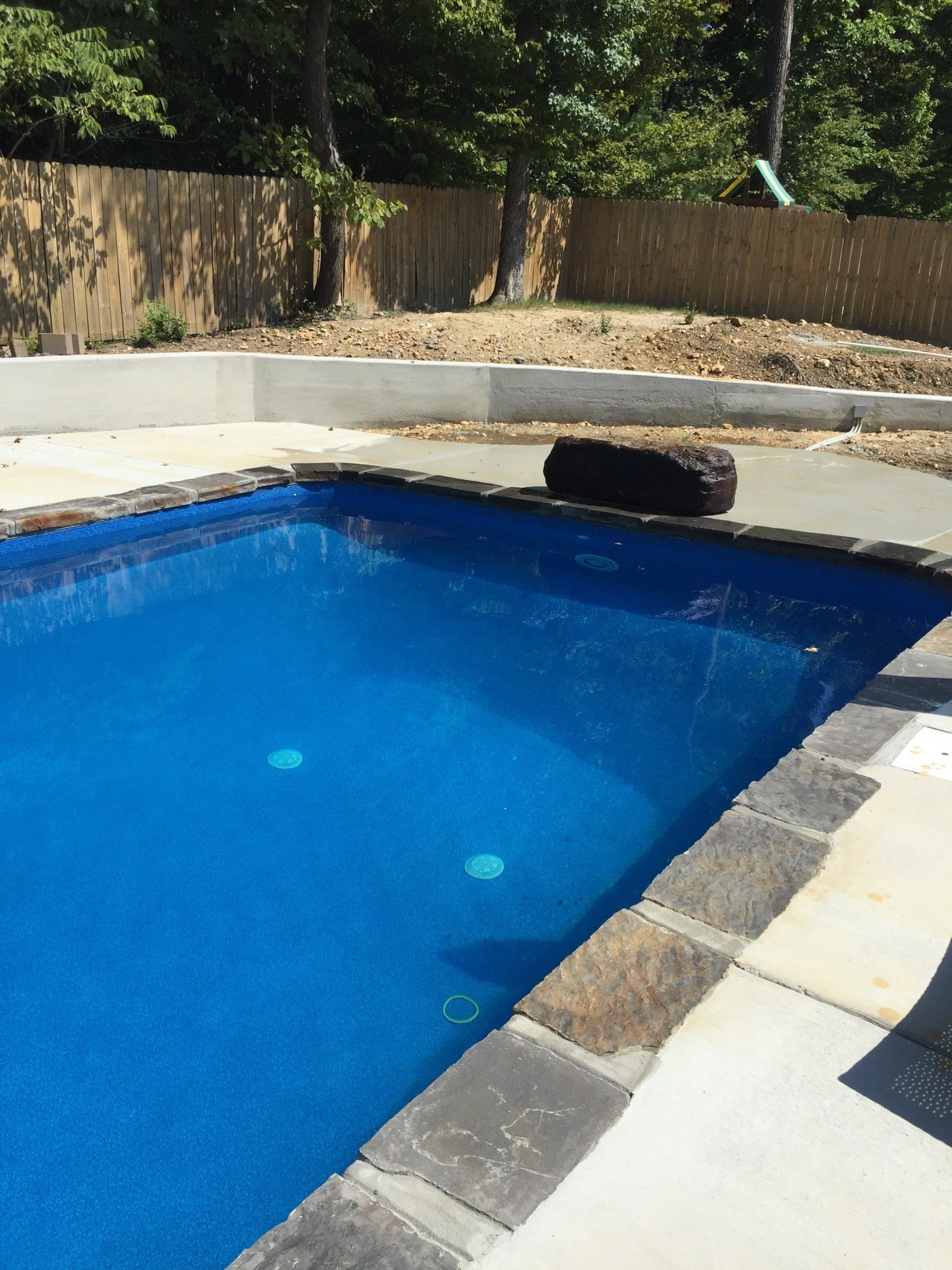 Blue swimming pool with stone deck, concrete wall, and wooden fence in a backyard.
